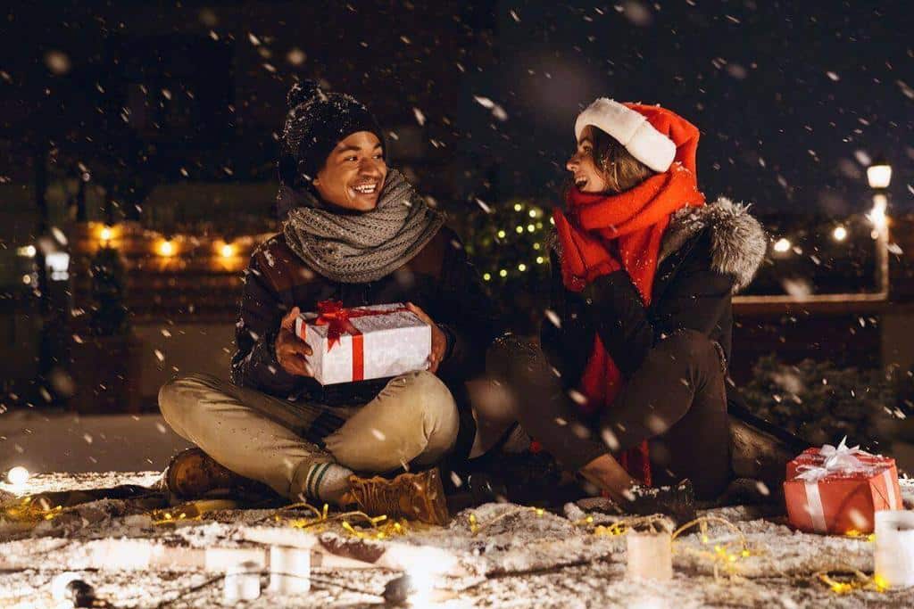 Een gelukkig paar in winterkleding zit buiten in de sneeuw, uitwisselend kerstcadeaus, omringd door feestelijke lichtjes en vallende sneeuwvlokken.