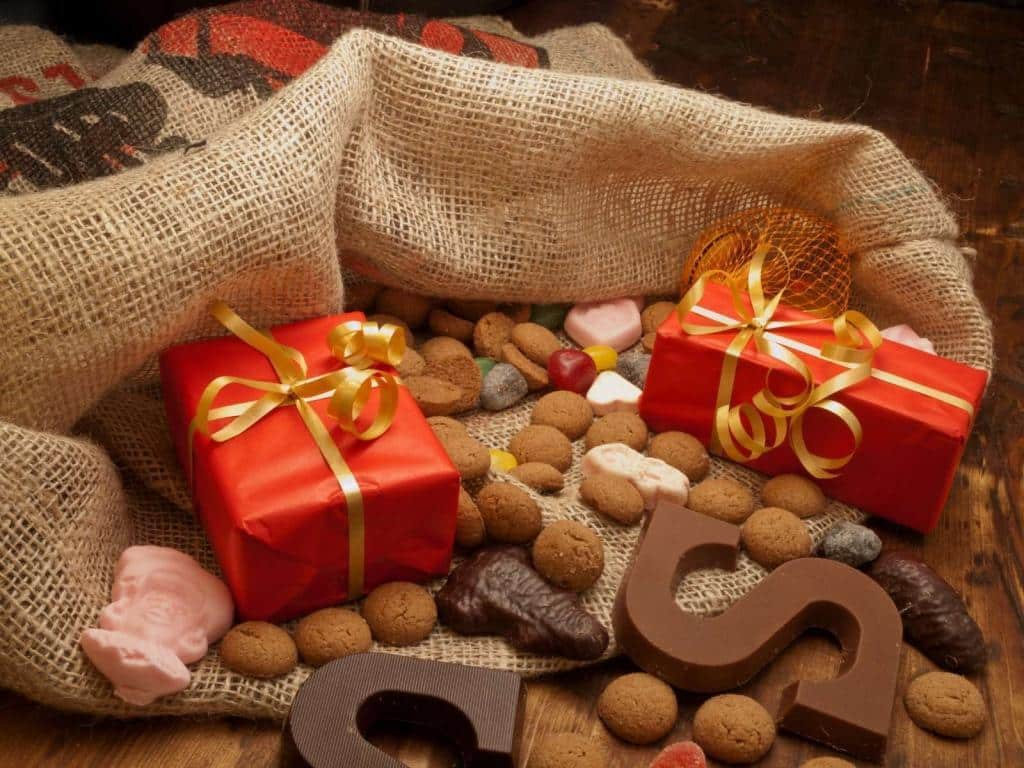 Sinterklaascadeau tussen de pepernoten, chocoladeletters en pieten zakken. De cadeaus zijn ingepakt met rood papier en een gouden lint.