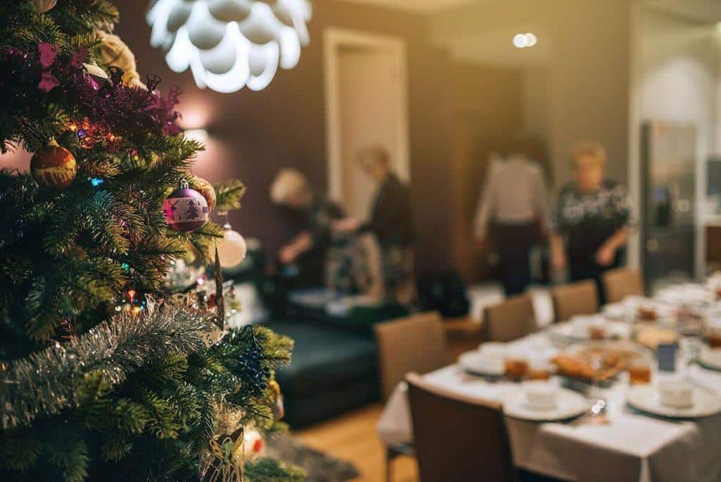Een feestelijk versierde kerstboom staat op de voorgrond met een onscherpe achtergrond van mensen die zich voorbereiden op een kerstdiner in een huiselijke setting.