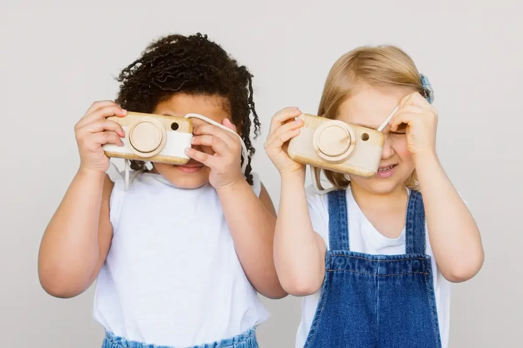 2 meisjes met een speelgoedcamera. Eén meisje heeft donker haar, de ander heeft blond haar en een tuinbroek.